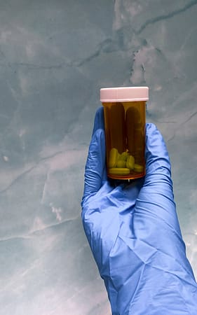 our-experience Close-up of a gloved hand holding a prescription pill bottle against a textured background.