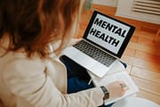 pexels-photo-3958396-3958396 A woman participating in an online therapy session, taking notes with a laptop displaying 'Mental Health'.