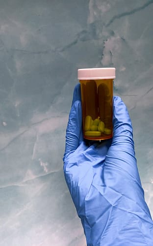 gallery-1 Close-up of a gloved hand holding a prescription pill bottle against a textured background.