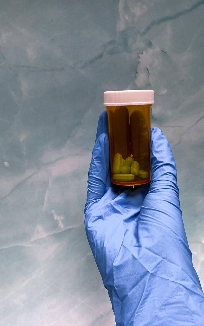 gallery-1 Close-up of a gloved hand holding a prescription pill bottle against a textured background.